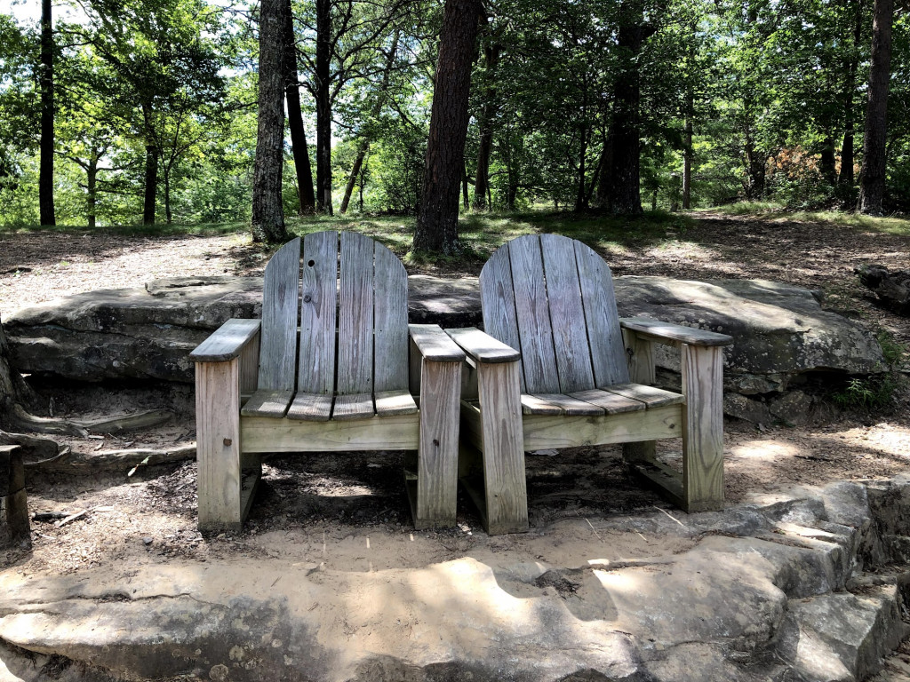 Two large wooden chairs sit side-by-side in a forest clearing.